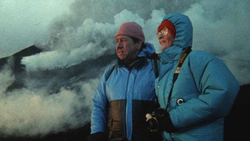 White middle-aged heterosexual couple stand on volcanic mountain in blue puffer jackets and beanies surrounded by smoke.