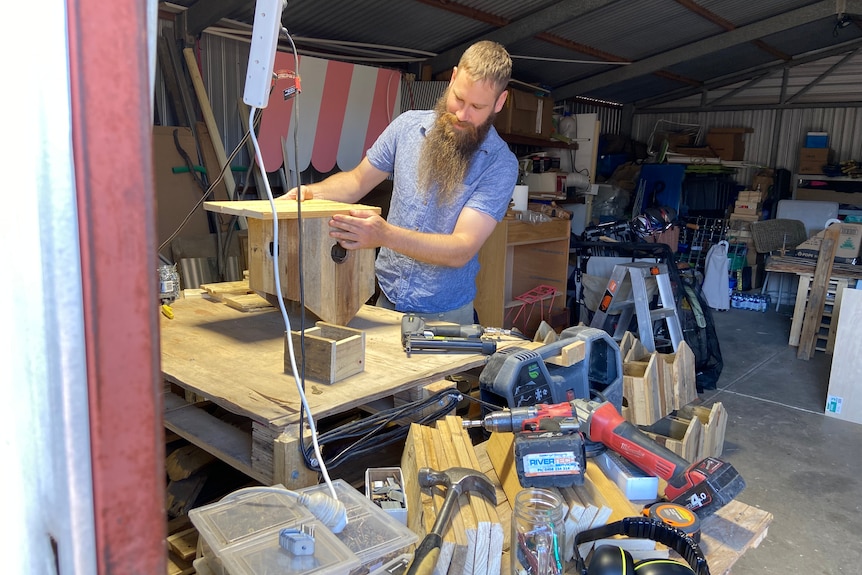 Man stands at bench with birdhouse in shed with work tools in foreground.