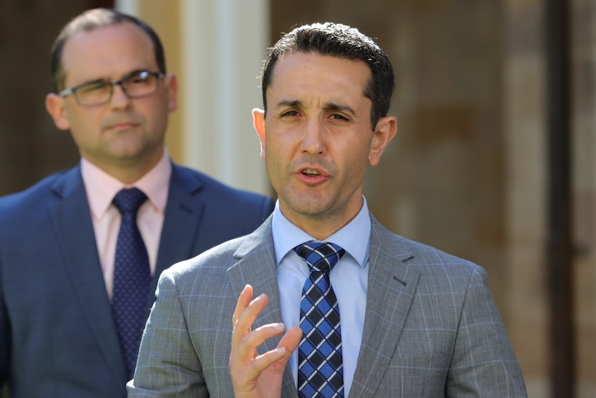 A man in a grey suit gestures as he speaks at a media conference. There is a bespectacled man in a blue suit behind him.