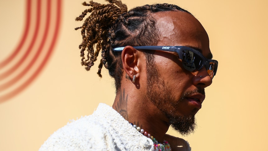Lewis Hamilton, wearing sunglasses and a wool shirt, walking though an F1 paddock in the afternoon.
