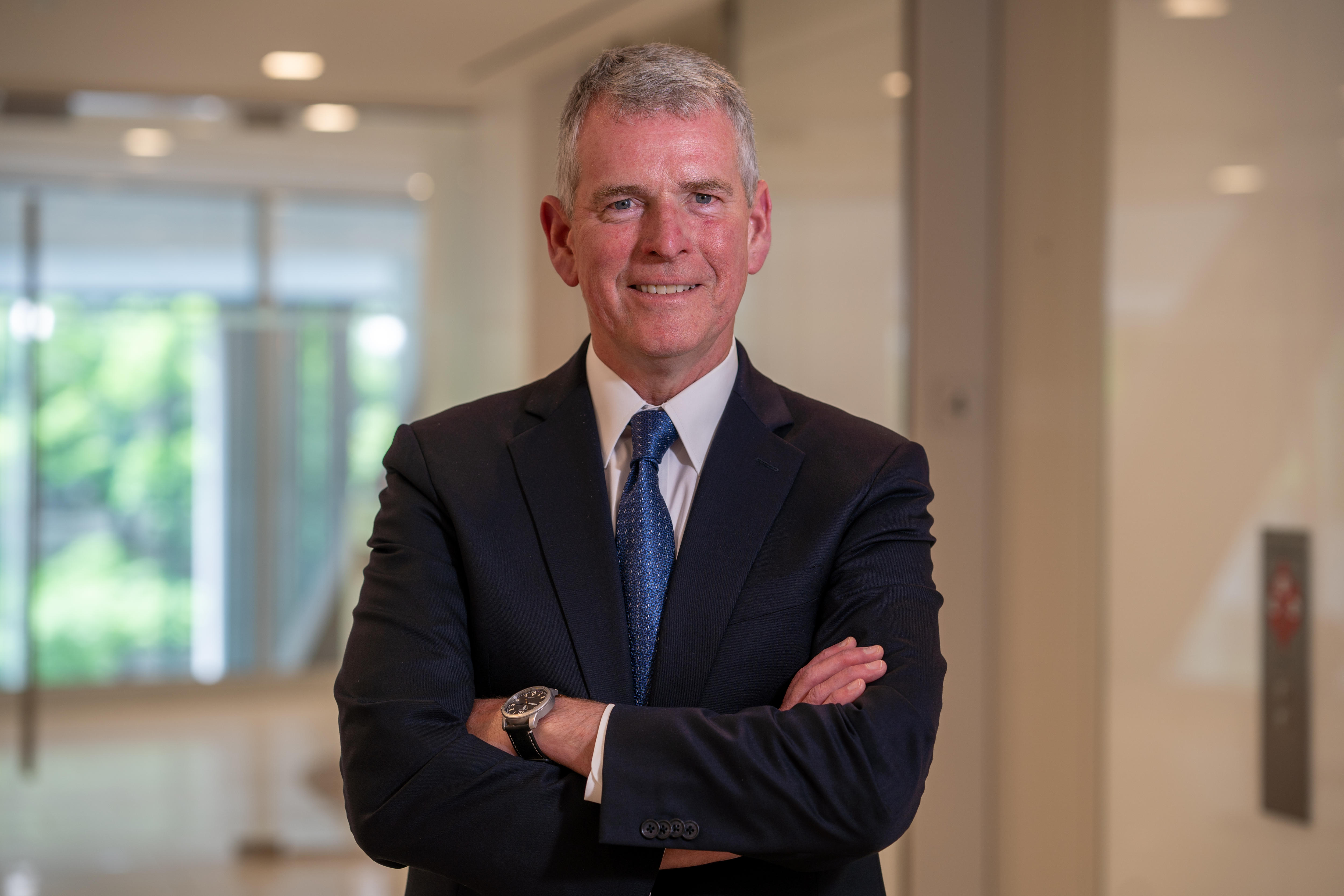 An older man with grey hair stands with arms folded across his chest smiling. He wears a dark suit over blue tie and white shirt