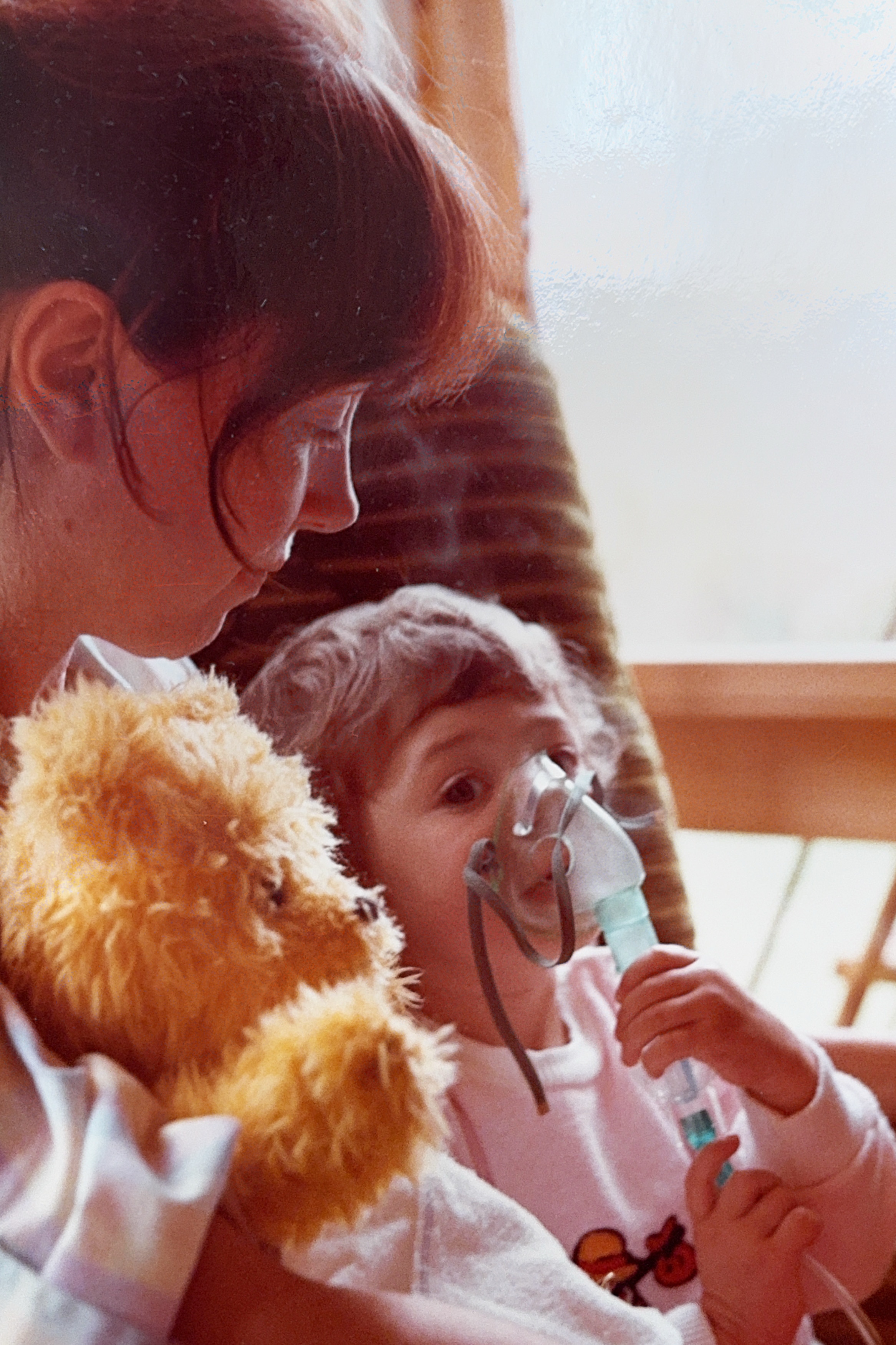  A woman holds a toddler in her arms. The toddler has an oxygen mask on and holds a brown teddy bear