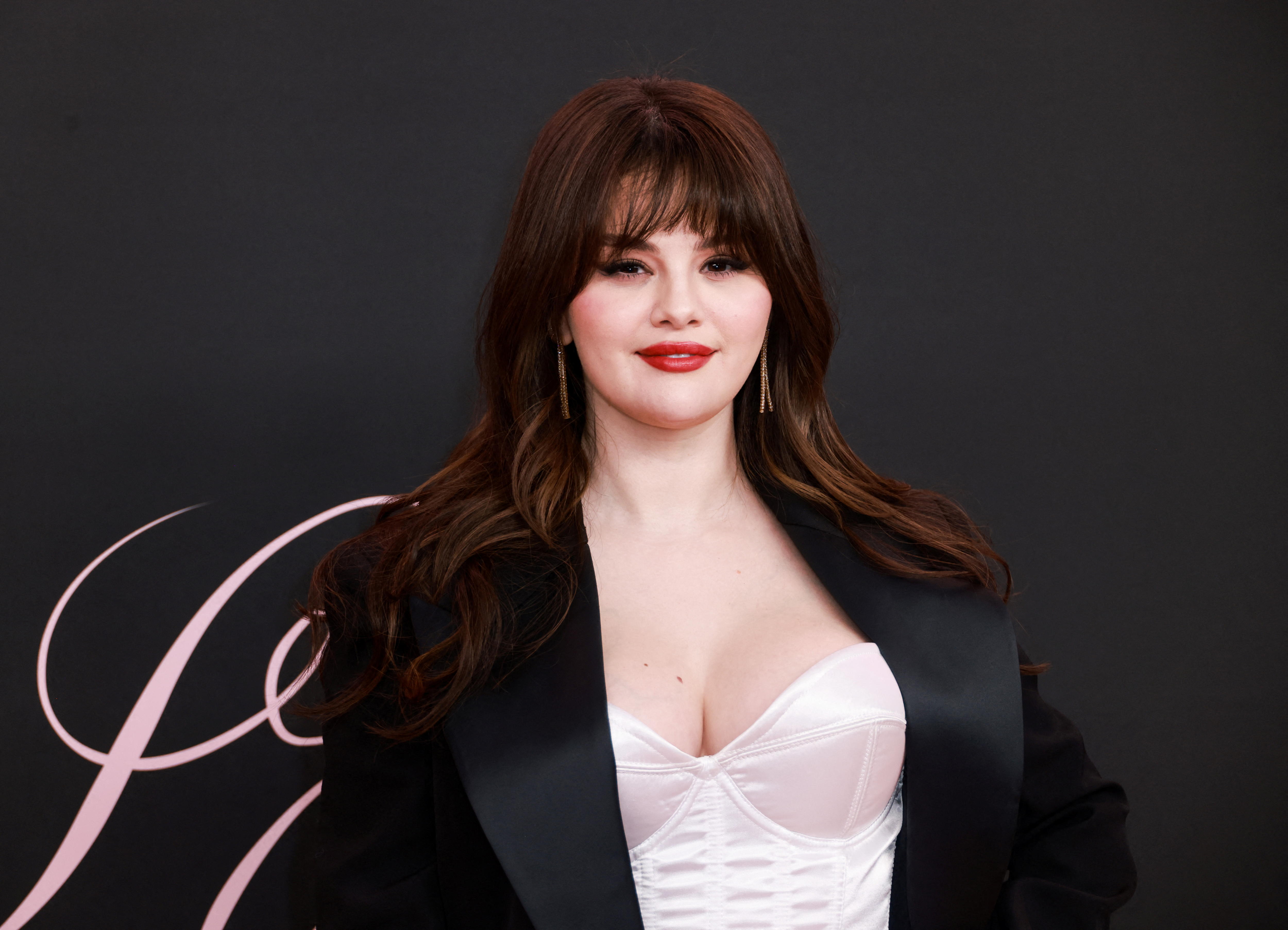 Selena Gomez con corpiño rosa y chaqueta de traje negra, lápiz labial rojo y cabello largo y oscuro