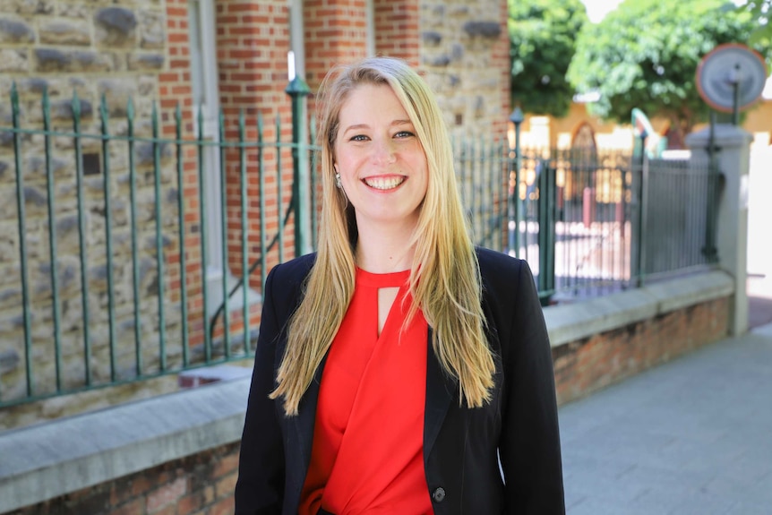 Woman with long, blonde hair standing in front of a fence around a red brick building. She's wearing a red top and black jacket.