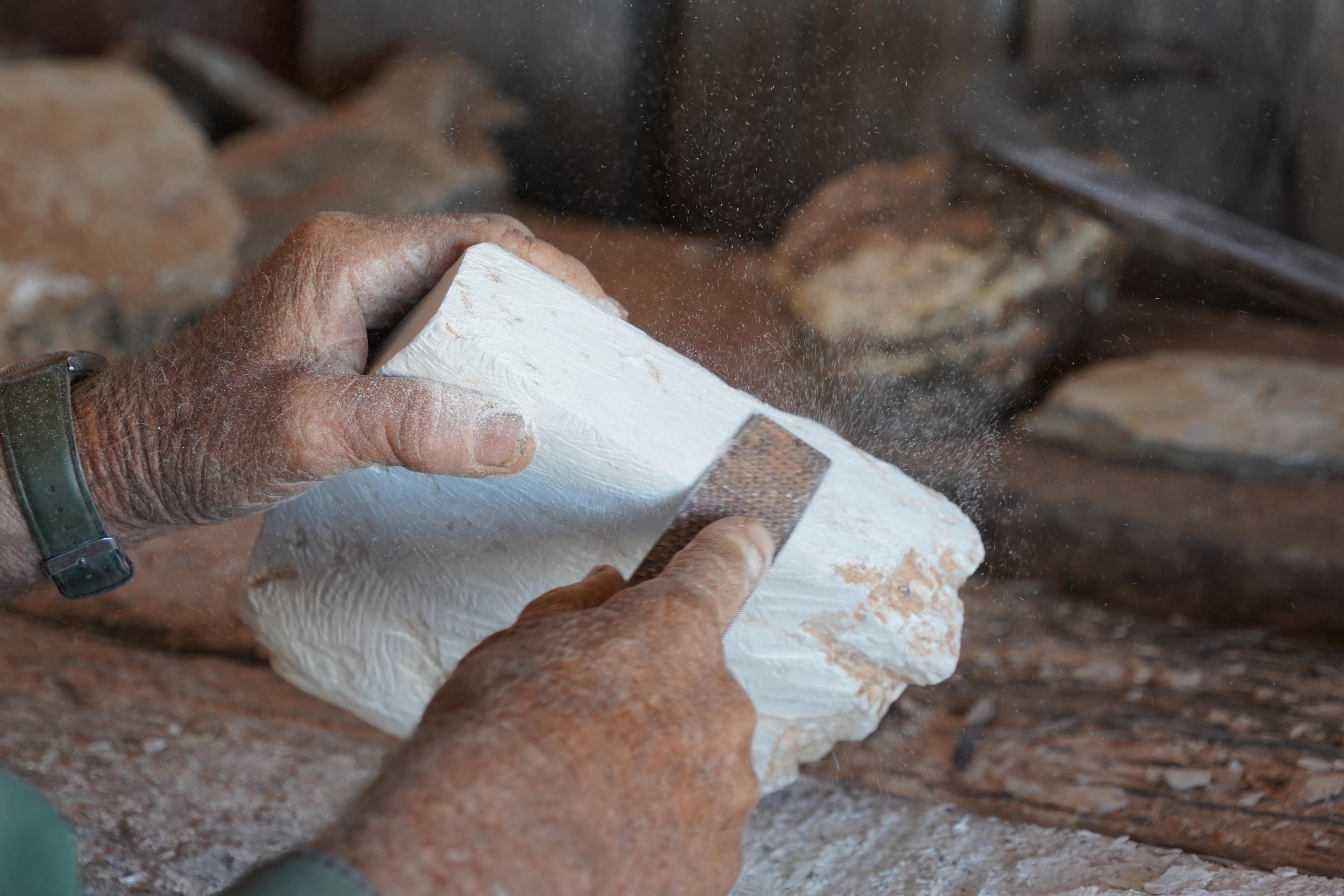 Primeros planos de una mano sosteniendo un trozo de piedra blanca y otra mano sosteniendo la escofina que raspa la piedra. 