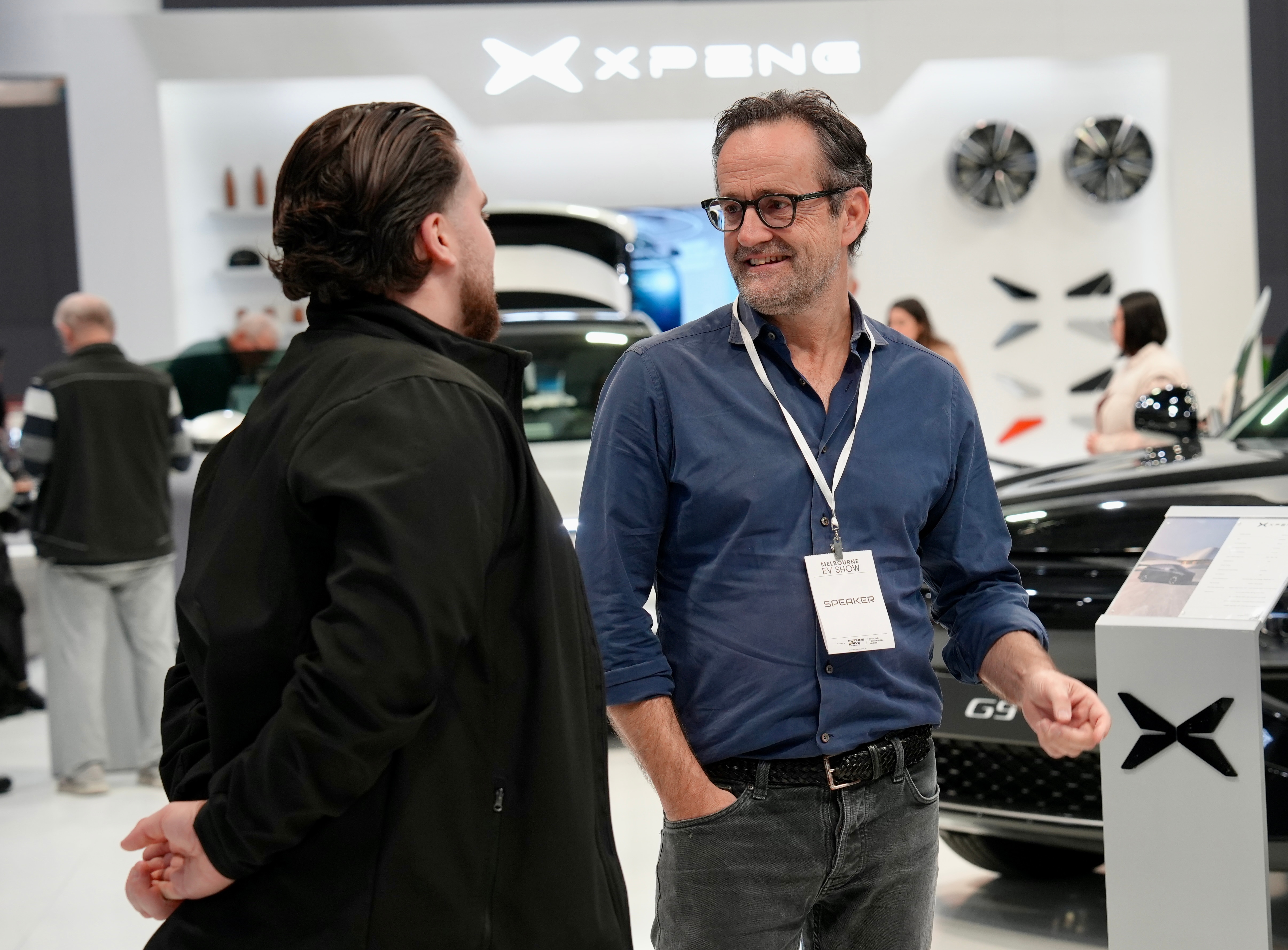 A smiling man with a pass hanging around his neck talks to a man in a black jacket.