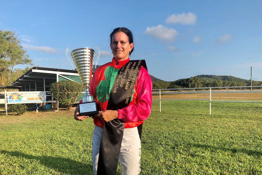 A diminutive female jockey in racing outfit proudly holds her trophy
