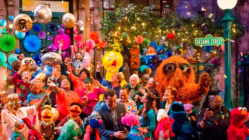 A group shot of the Sesame Street show set shows the cast looking up at confetti and balloons.