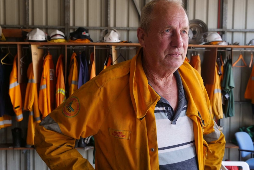 Hank Rappard stands in a shed, with firefighter protective clothing and uniforms hanging behind him.