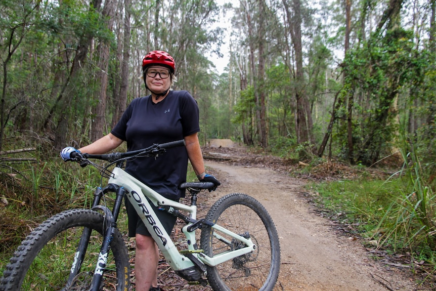 a woman stands beside a mountain bike. Behind her is a wooden mountain bike trick