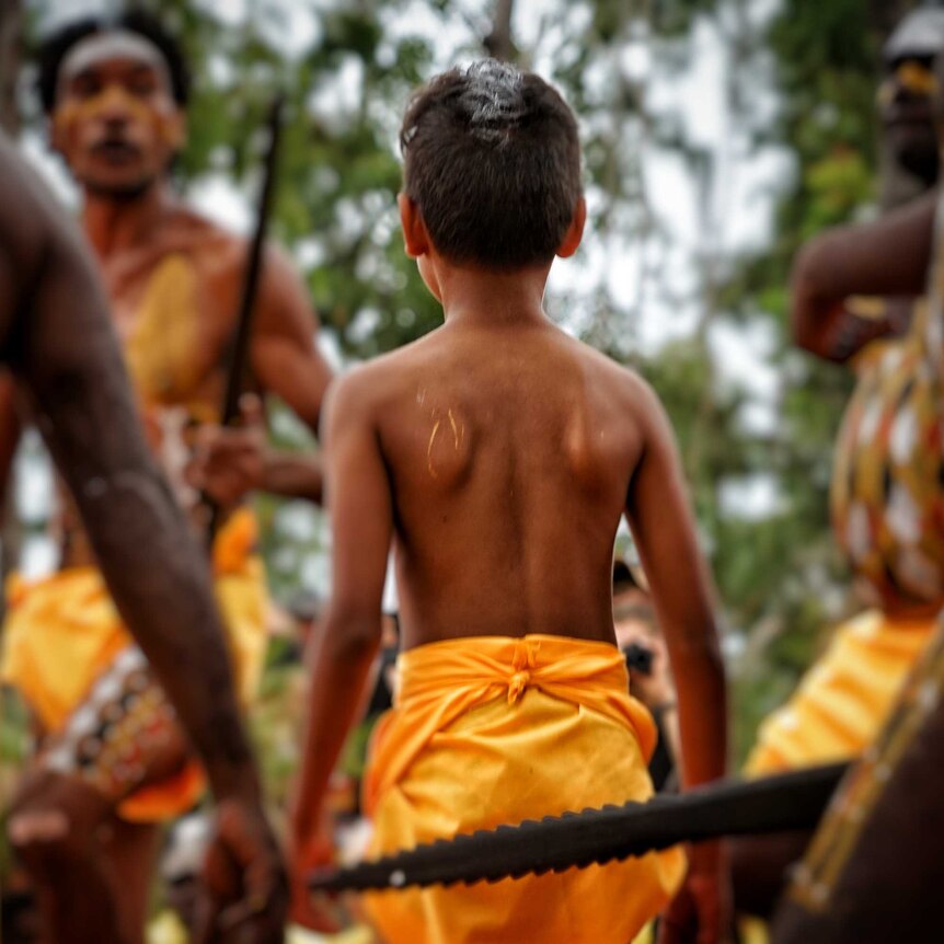 A young boy in a traditional outfit stands among men with spears at Garma festival.