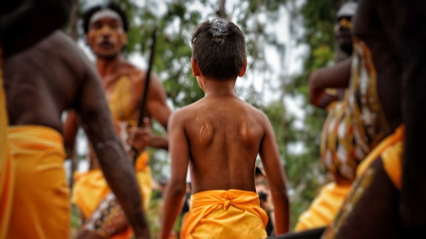 A young boy in a traditional outfit stands among men with spears at Garma festival.