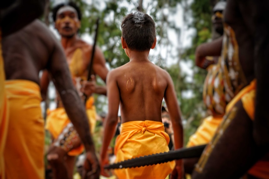 A young boy in a traditional outfit stands among men with spears at Garma festival.