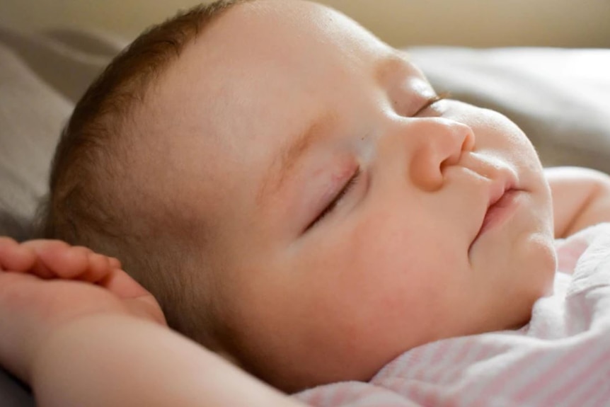 Close-up shot of young baby with hands up by head peacefully asleep with mouth closed.