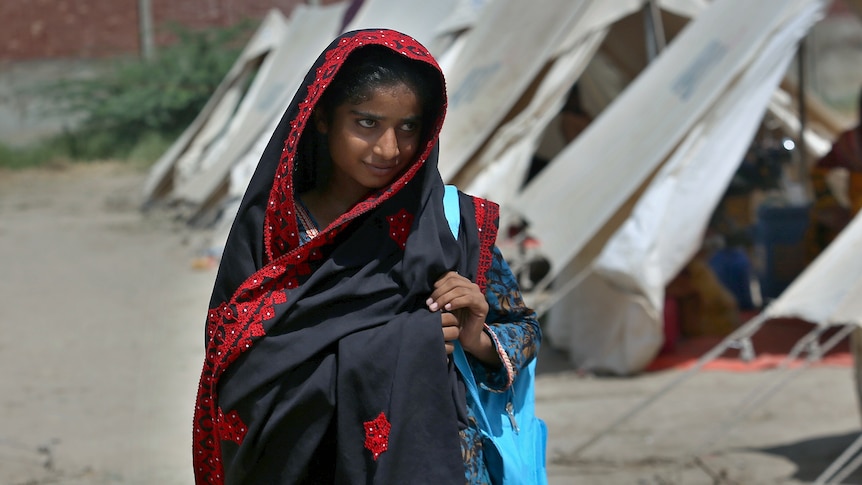 A Pakistani girl in a navy and red shawl carries a bag along a dusty path past a series of plain white tents.