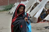 A Pakistani girl in a navy and red shawl carries a bag along a dusty path past a series of plain white tents.