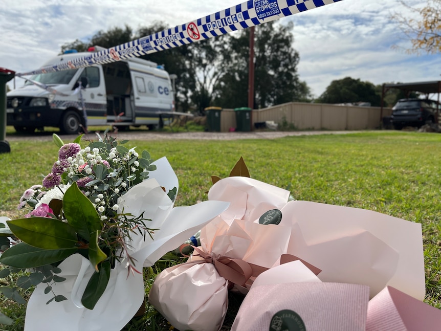 Flowers left outside a house with police tape above the flowers and a van in the background.