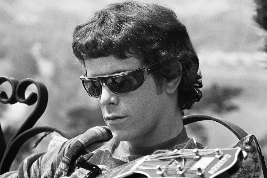 A black and white image of Lou Reed sitting on a bench wearing sunglasses as he plays guitar