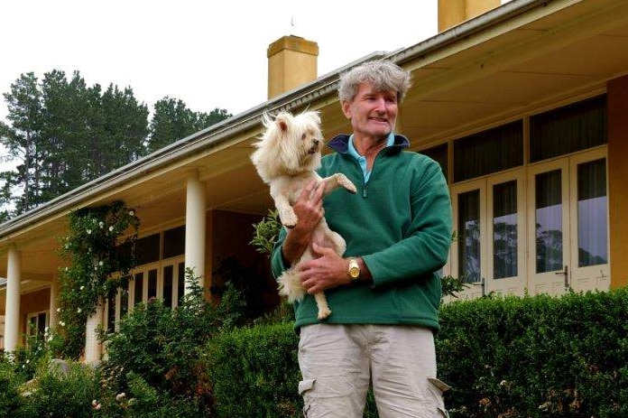 Middle aged white man holds well-groom dog in front of stately single-level home on rural property