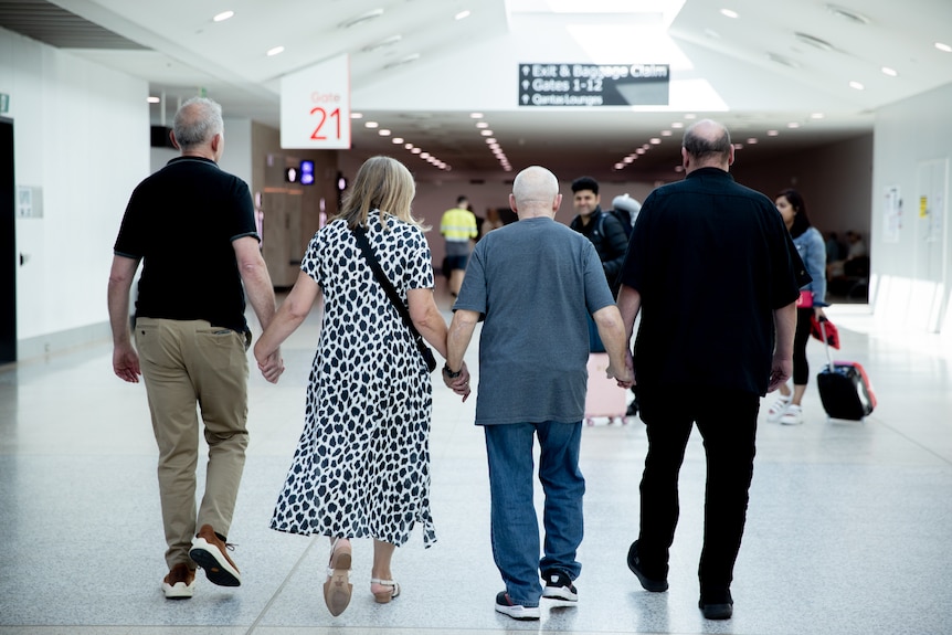 Four older people walk hand-in-hand through an airport with their backs facing the camera