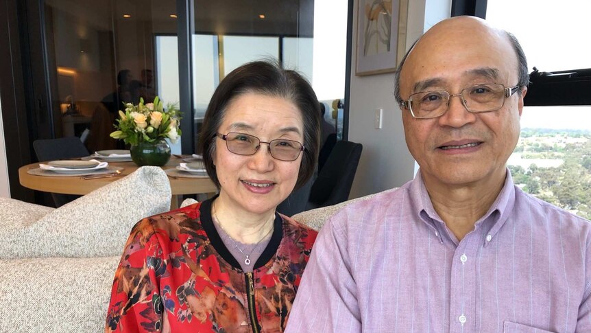 A Taiwanese woman with short black hair, glasses, red floral cardigan sits on couch next to bald man with glasses in pink shirt