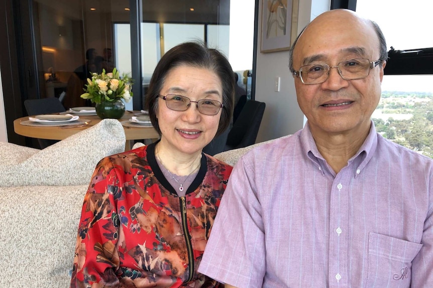 A Taiwanese woman with short black hair, glasses, red floral cardigan sits on couch next to bald man with glasses in pink shirt