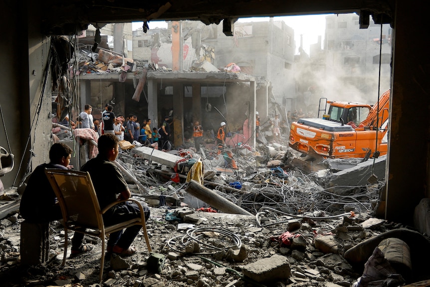 Two men sit on chairs and look out to a street filled with rubble and destroyed buildings