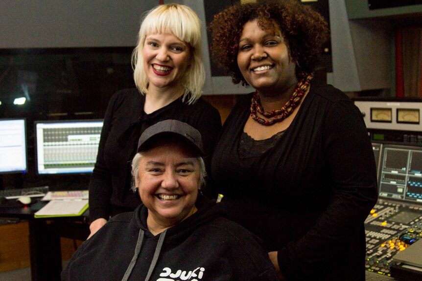Three women stand in the control room of a radio studio.