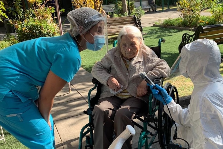 An elderly woman sits in a wheelchair, smiles, while two people wearing masks crouch down to talk to her. One holds a microphone