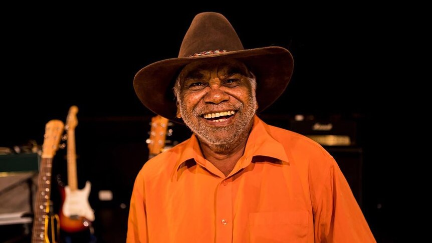 Warren H Williams wears an orange shirt, brown hat and he has a huge smile on his face. The are guitars in the studio behind.