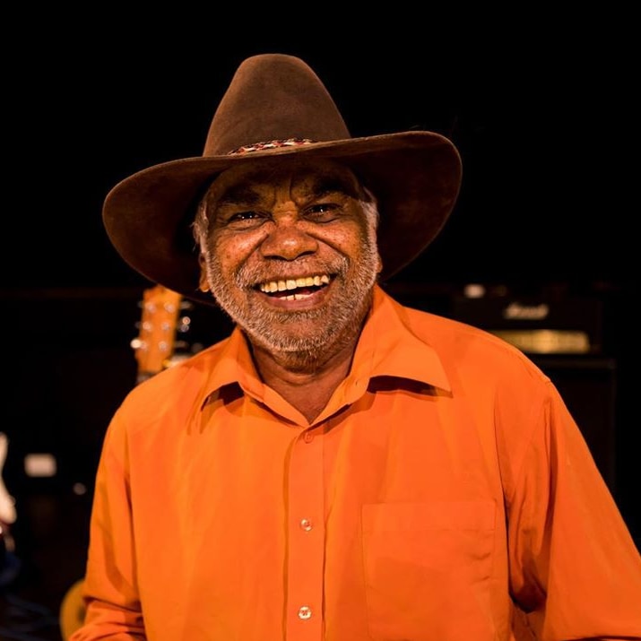 Warren H Williams wears an orange shirt, brown hat and he has a huge smile on his face. The are guitars in the studio behind.