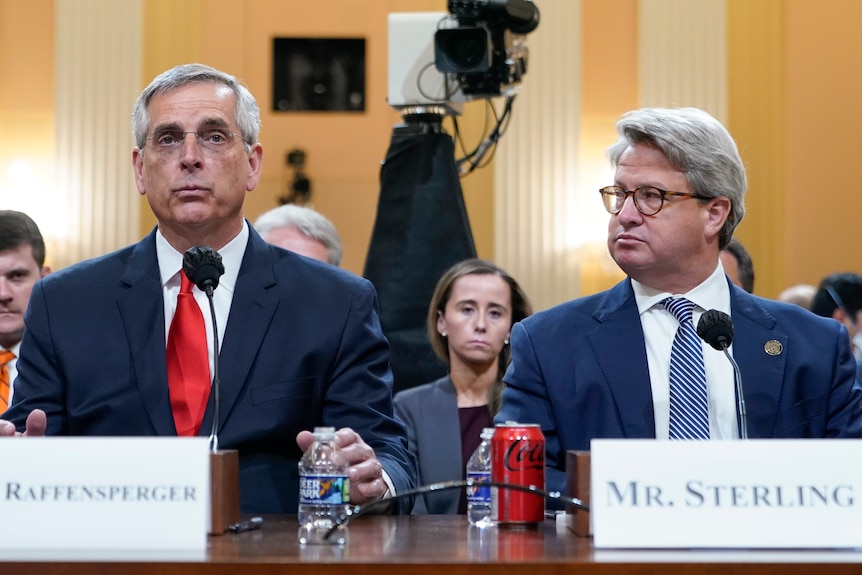 Brad Raffensperger in a red tie speaks at a desk while his deputy Gabe Sterling looks on