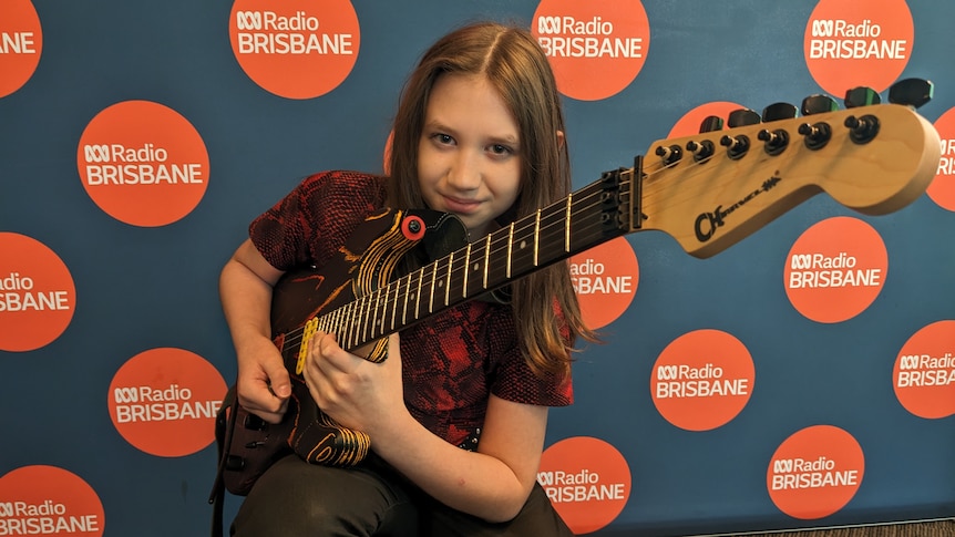 A smiling 12-year-old girl with long, loose hair squats as she holds a dynamic electric guitar.