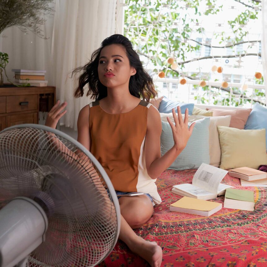 A woman sits on her bed fanning herself and sitting in front of a pedestal fan