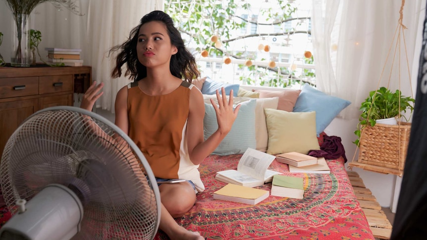 A woman sits on her bed fanning herself and sitting in front of a pedestal fan