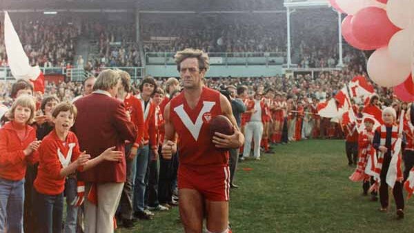 A veteran SANFL footballer runs out on an oval with a football under his arm, as fans cheer and hold red and white balloons.