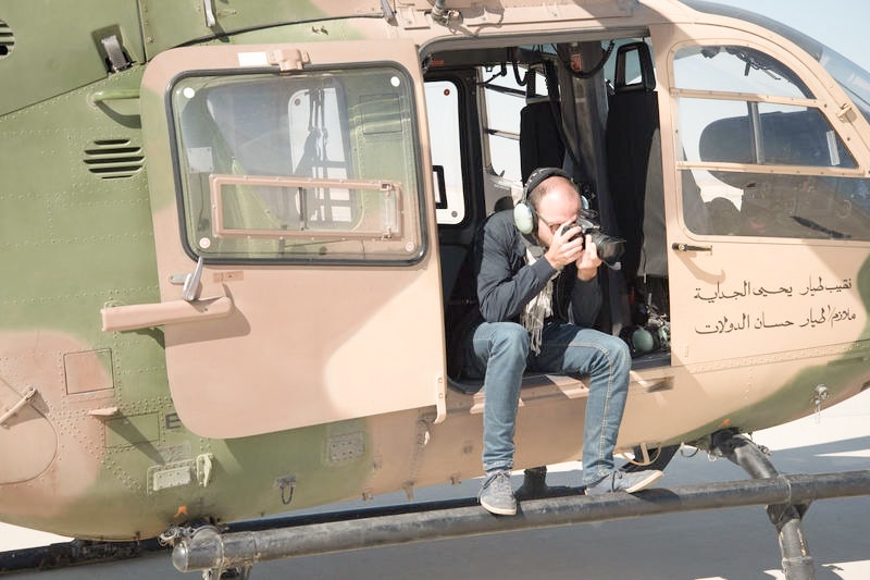 A man sits on the edge of a helicopter with a camera taking photos of the sky.