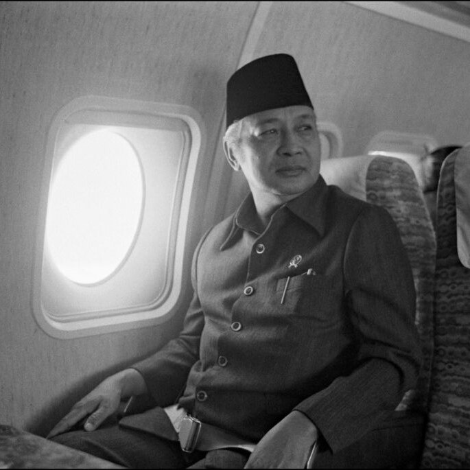 Suharto sits on a plane wearing a hat gazing right of frame