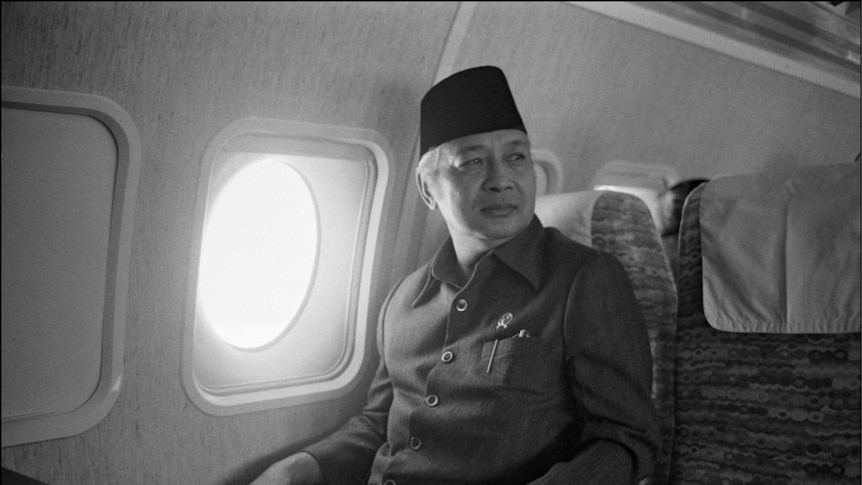 Suharto sits on a plane wearing a hat gazing right of frame