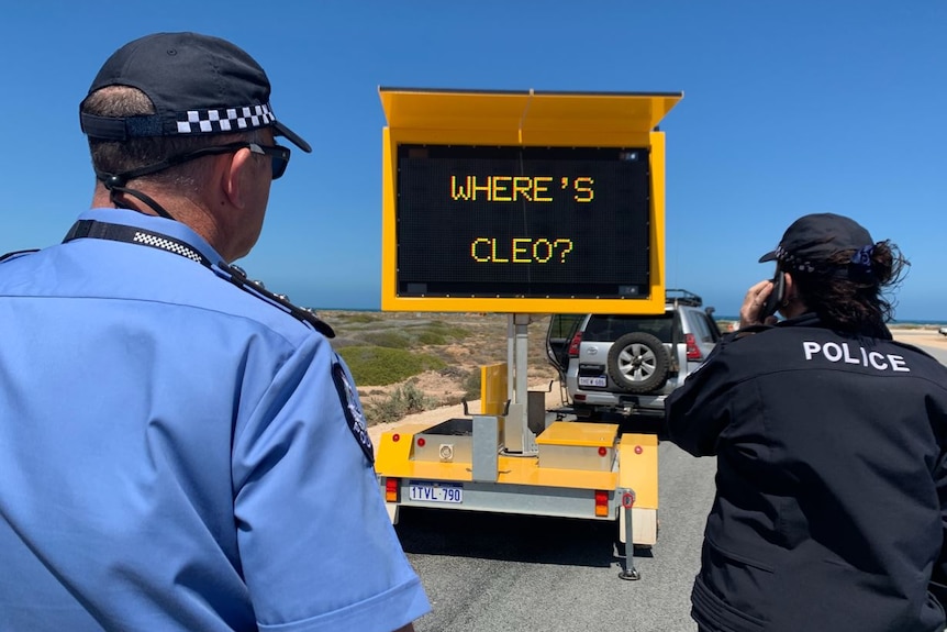 Police pictured from behind as they look at a sign on the side of the road says: 'Where's Cleo?'