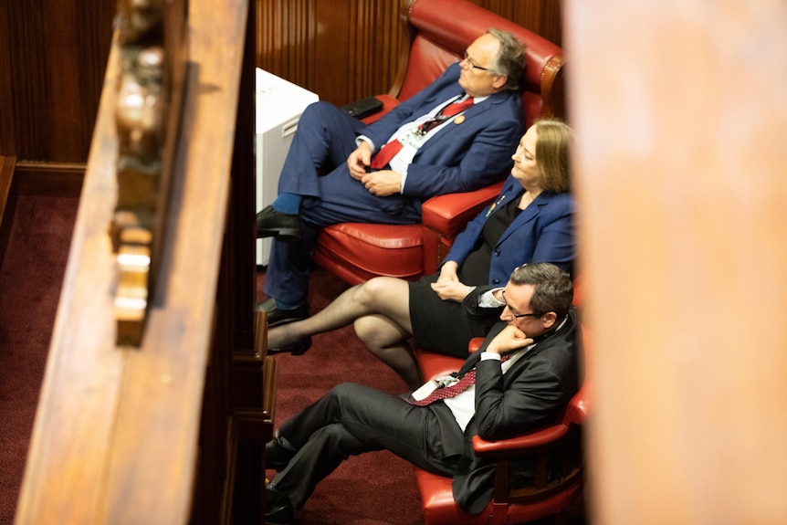 A man in a blue suit, a woman in a blue jacket and a man in a black suit look to the left watching something.