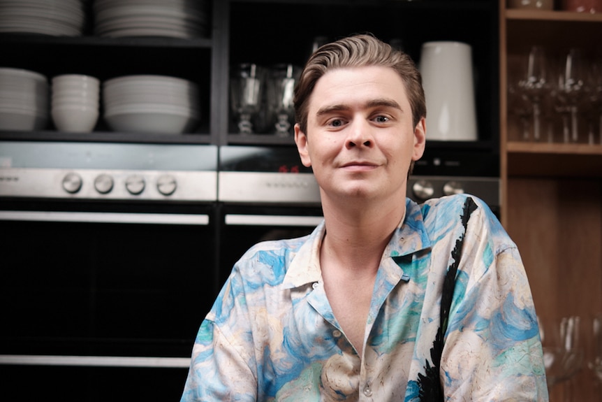 A young man leans on the kitchen counter and smiles slightly.  There is a baking tray in front of him.