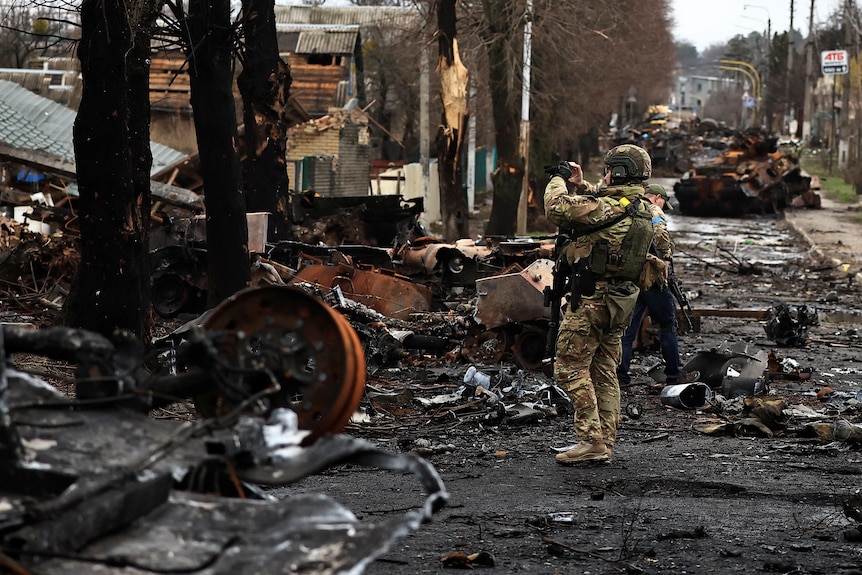 A soldier in military fatigues video tapes damage in war zone in scarred and blackened city street