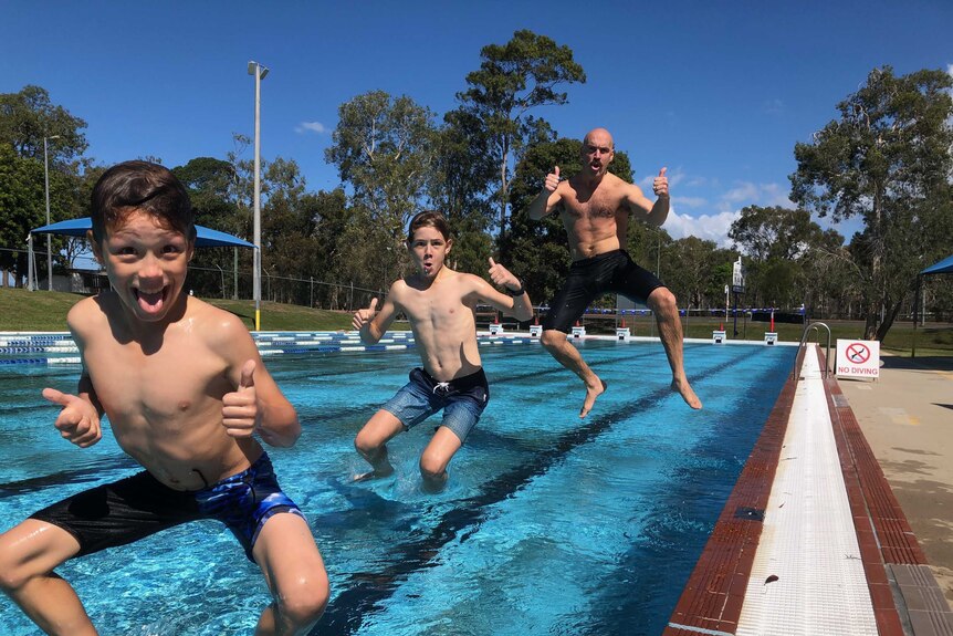 Two young boys and a man leapfrog into a pool at the same time, giving their thumbs up.