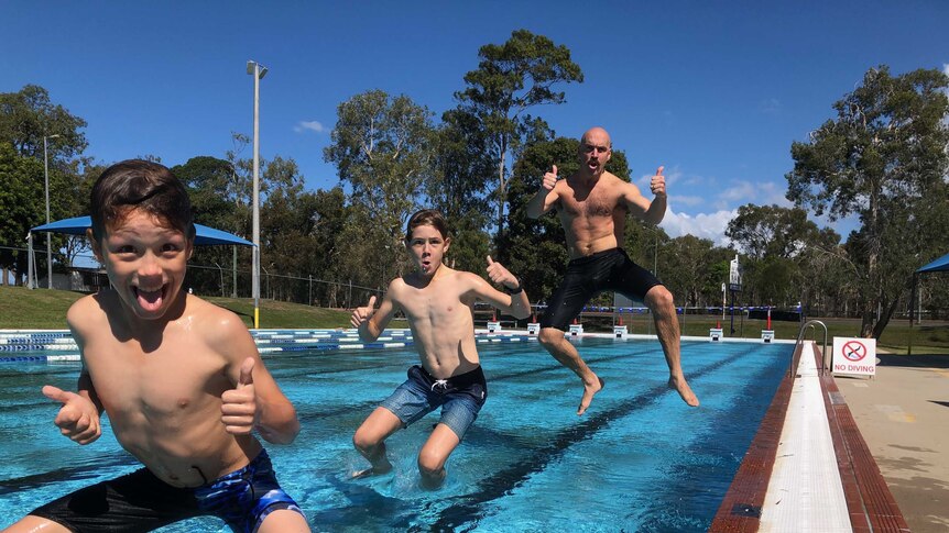 Two young boys and a man leapfrog into a pool at the same time, giving their thumbs up.