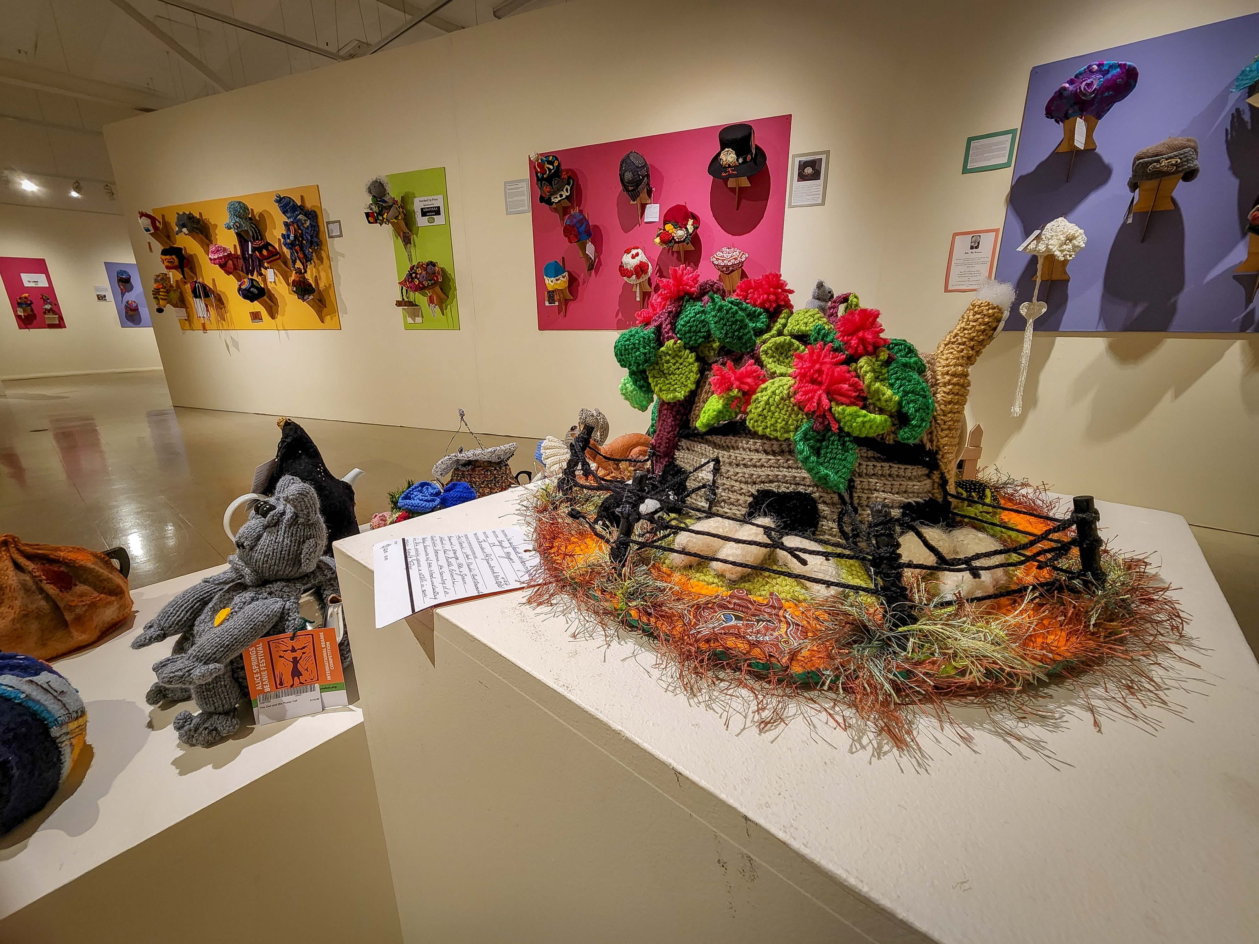 Knitted beanies on display inside a gallery, one in the foreground depicts a farmyard scene featuring tiny woollen sheep.