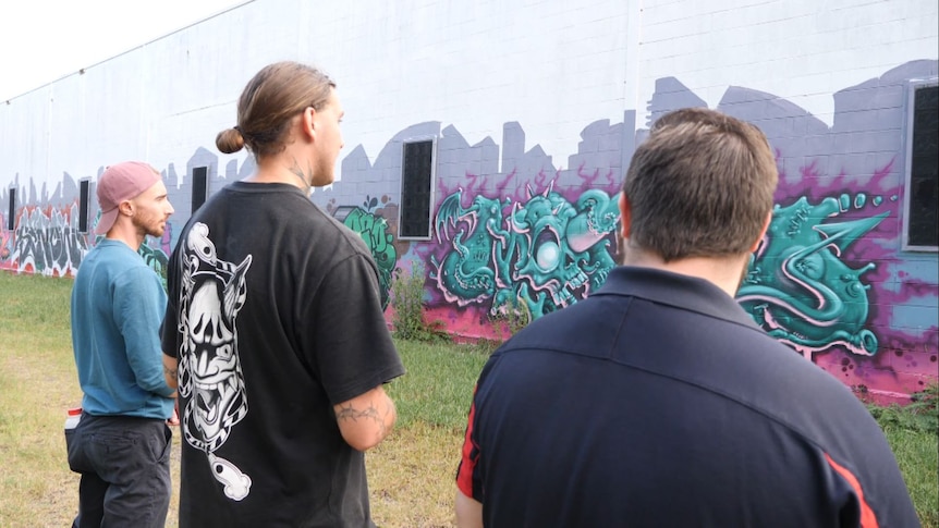 Three men stand in front of a graffiti wall with large colourful artworks.