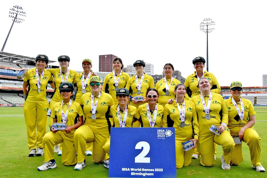 A team photo featuring two rows of female Australian blind cricketers smiling and posing with silver medals.