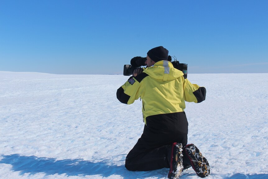 Peter kneels on the snow while looking into a camera which is resting on his shoulder.