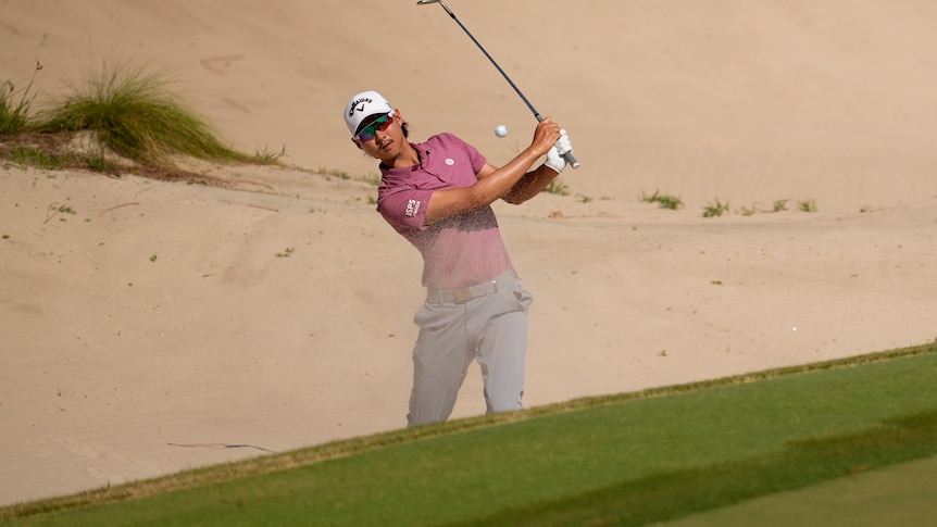Min Woo Lee plays a shot out of a bunker, with the ball seen between his face and the golf club